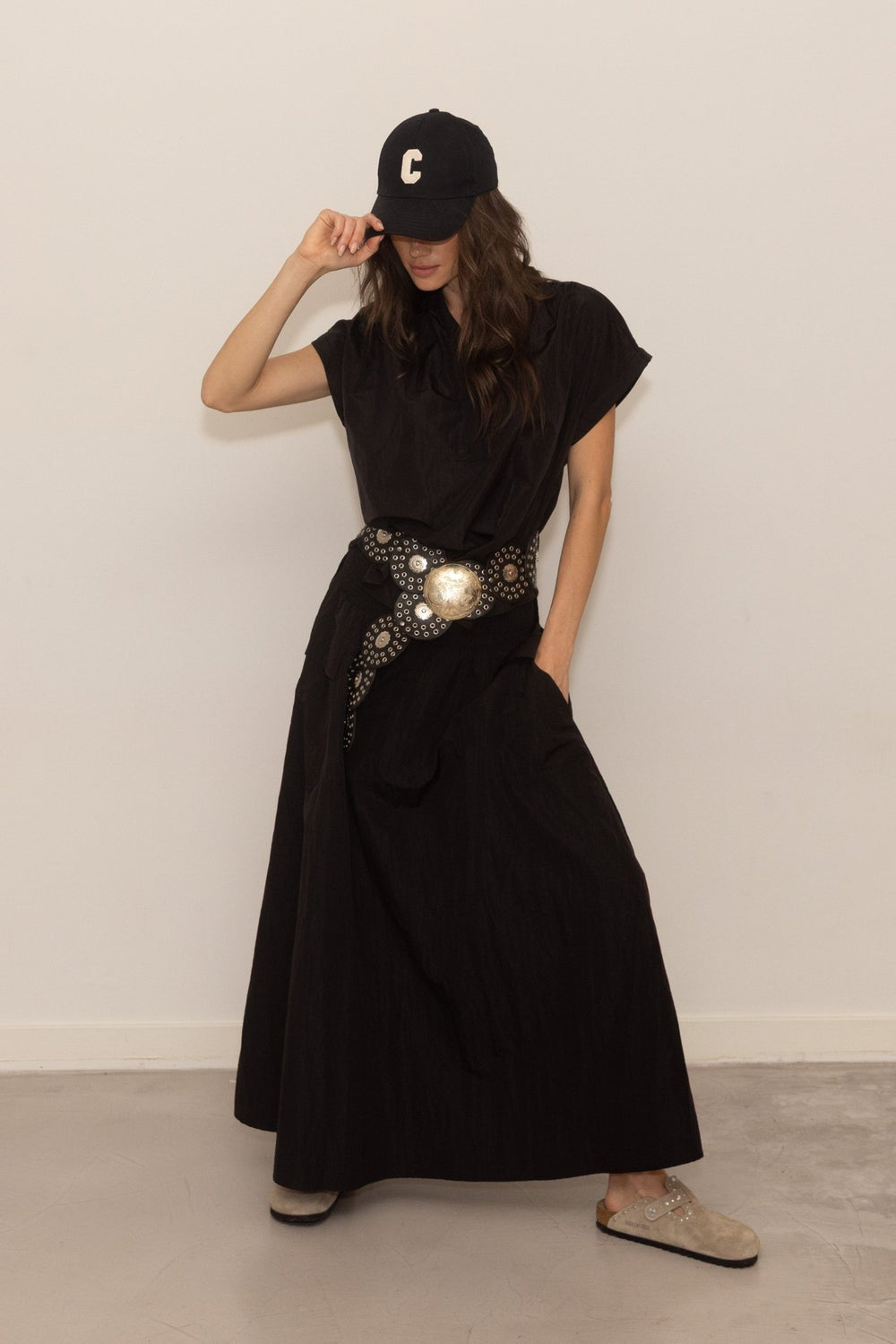 Woman wearing a black maxi skirt with a decorative belt and cap, standing against a plain background.