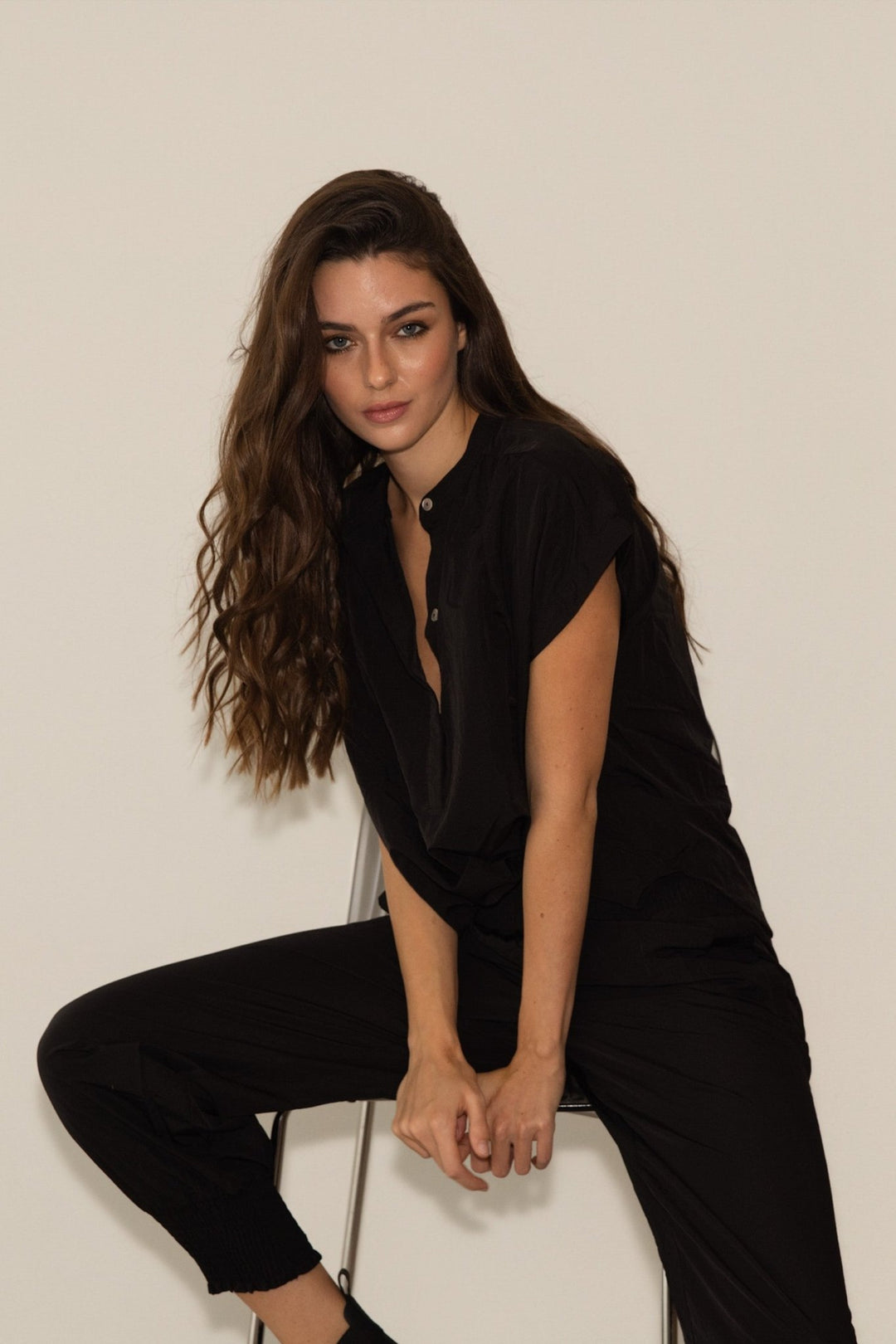 Woman in a black outfit sitting on a white stool against a plain background