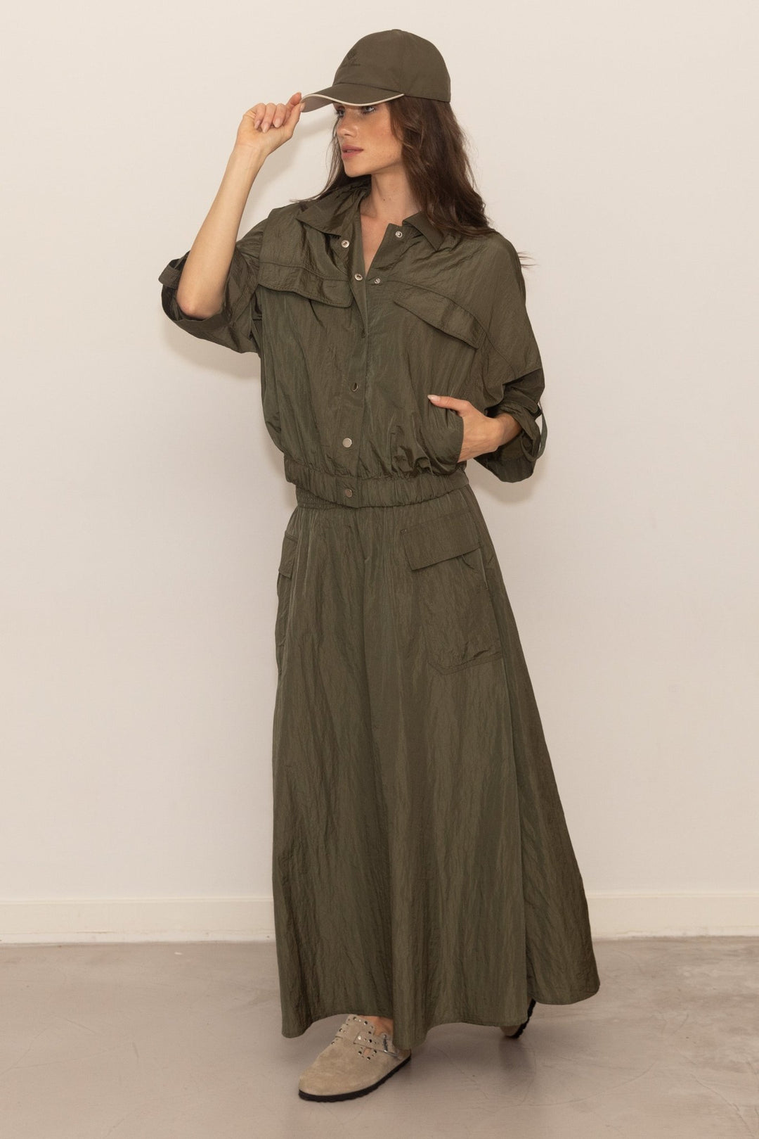 Woman wearing a long olive green jacket and pants with a cap against a white background