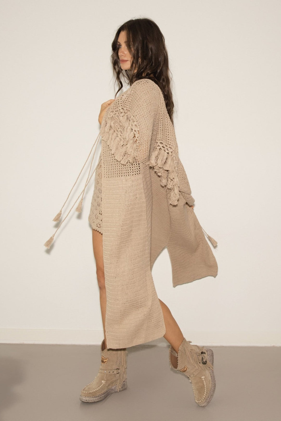 Woman wearing a beige crochet cardigan and dress with matching boots on a plain background