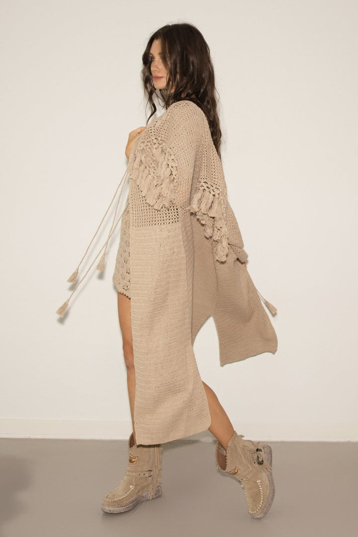 Woman wearing a beige crochet cardigan and dress with matching boots on a plain background