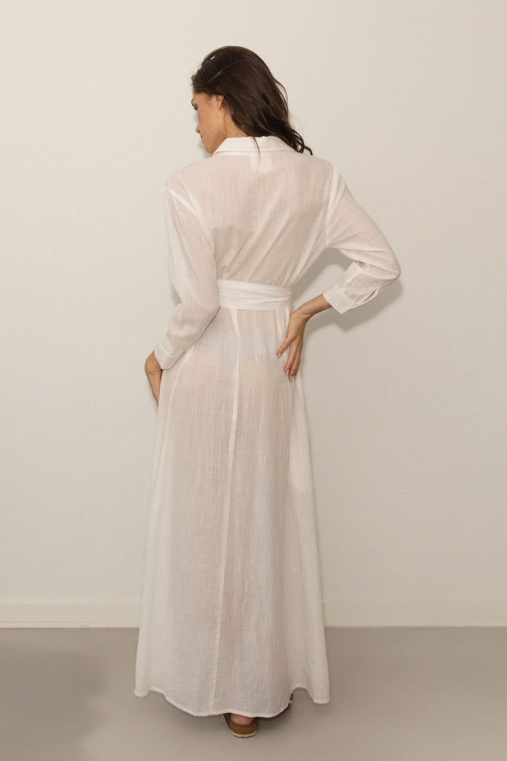 Woman wearing a long, light-colored dress with a belt against a plain background