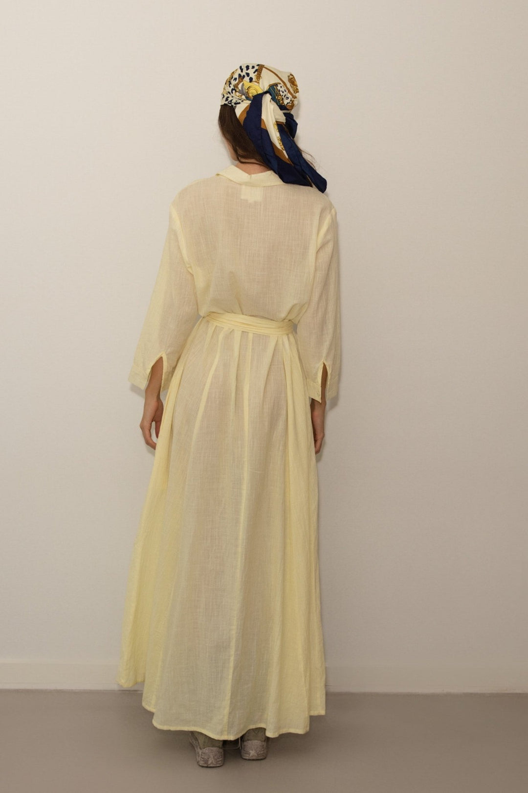 Woman wearing a long light yellow dress with a headscarf against a plain background