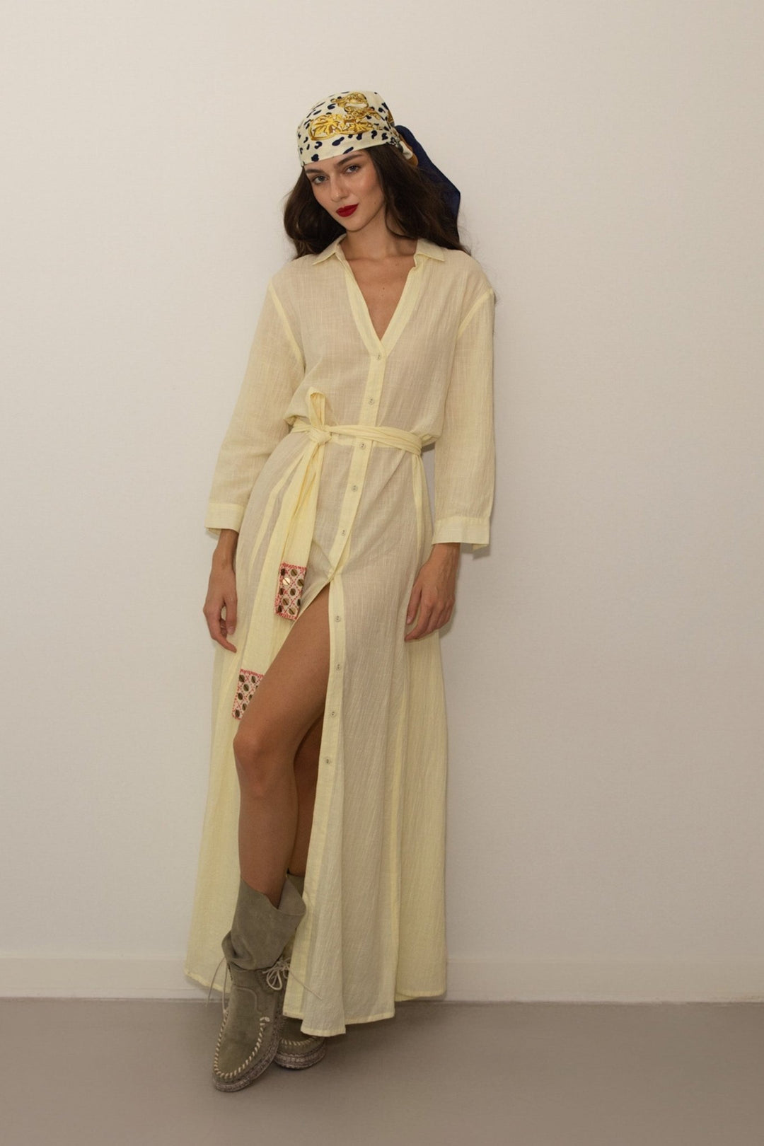 Woman wearing a long light yellow dress with a belt, standing against a plain background.