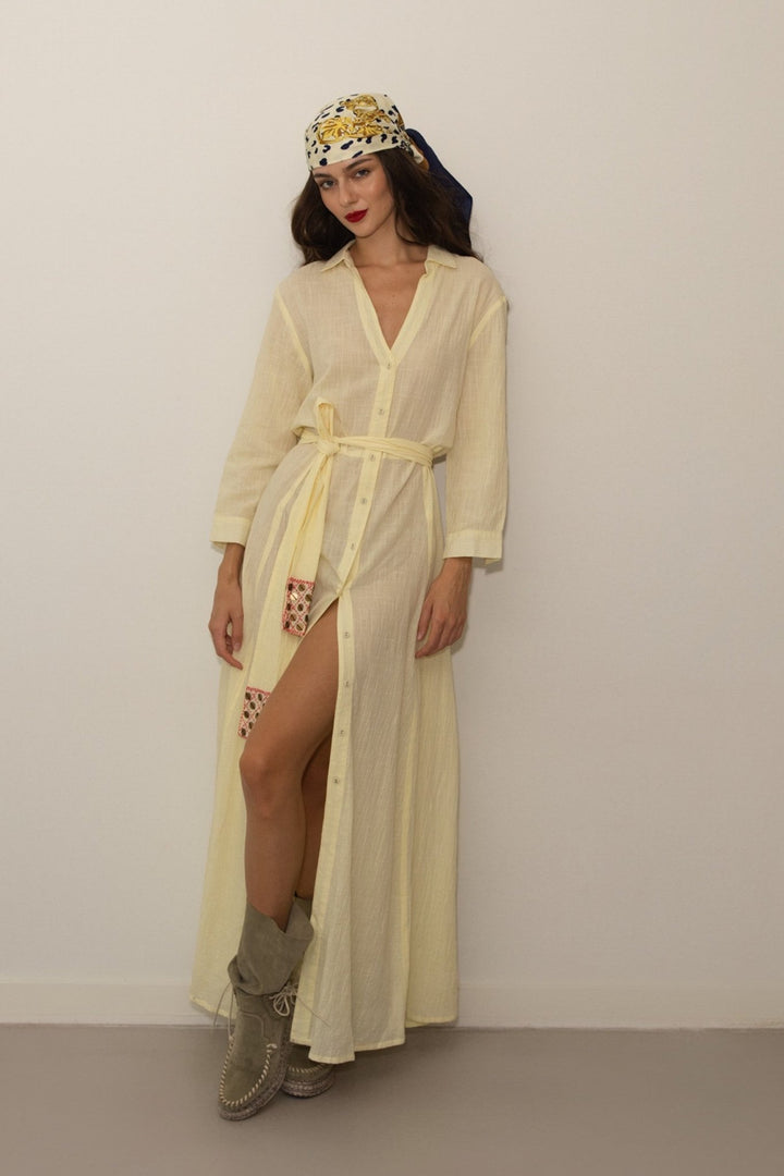 Woman wearing a long light yellow dress with a belt, standing against a plain background.