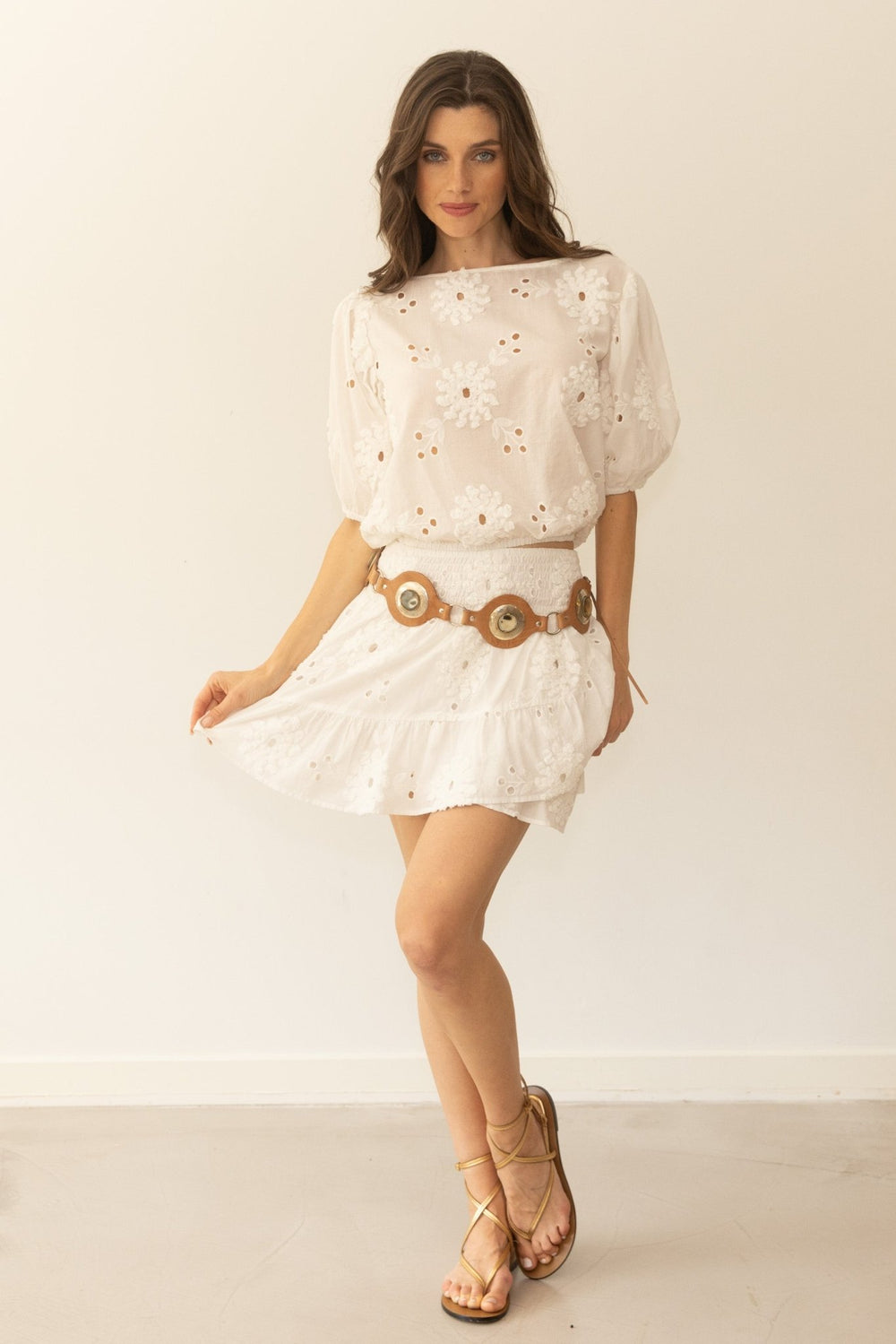 Woman wearing a white dress with floral details and a brown belt against a plain background